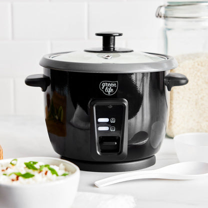 Black 6-cup rice cooker on a counter next to a bowl of cooked rice, a spoon, and some uncooked rice.