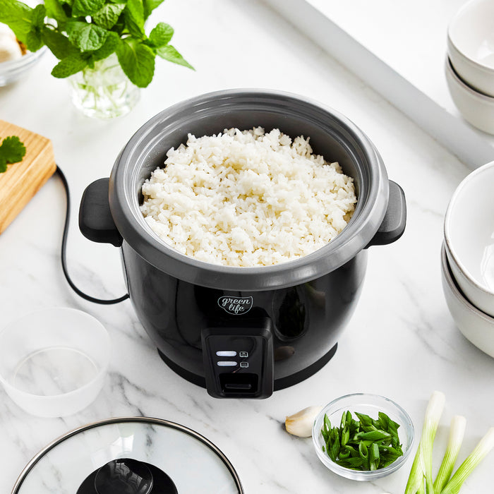 Black 6-cup rice cooker with ceramic coating on marble countertop, next to bowls and fresh herbs.