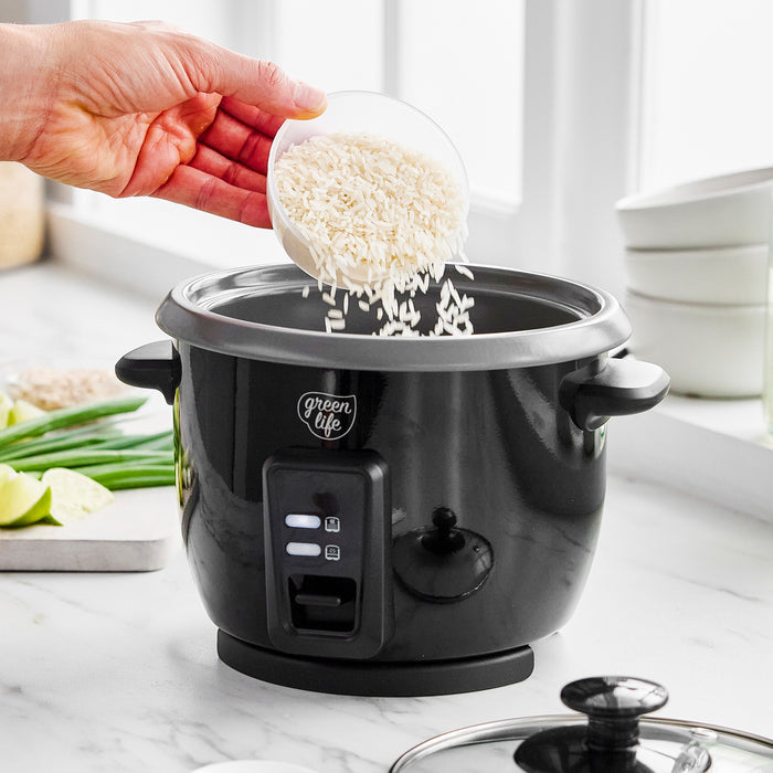 A hand pours rice into a black GreenLife 6-cup rice cooker on a white counter beside fresh vegetables.