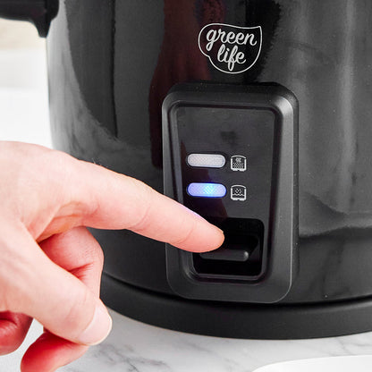 Hand pressing button on black 6-cup GreenLife rice cooker with ceramic coating and indicator lights.