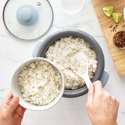 Cooked white rice being scooped from a blue GreenLife 6-cup rice cooker into a bowl on a white counter.
