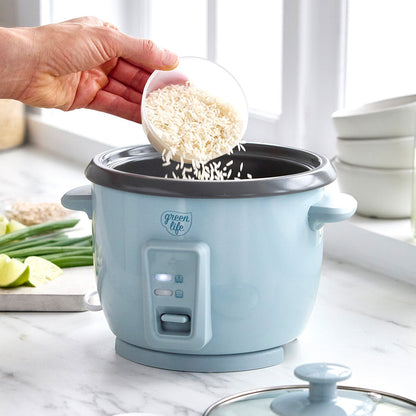 Uncooked rice being poured into a blue GreenLife 6-cup rice cooker on a kitchen counter.