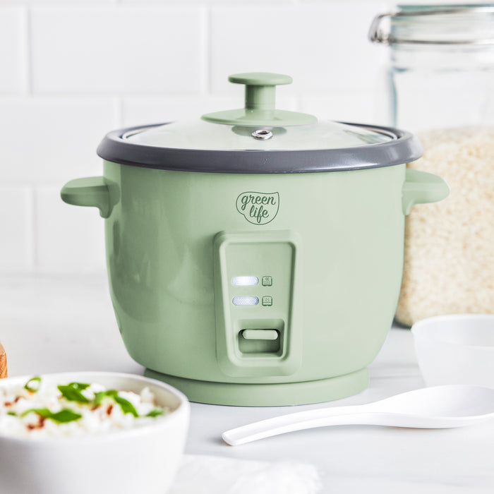 Sage green 6-cup rice cooker on a counter with a bowl of cooked rice, spoon, and a jar of uncooked rice.