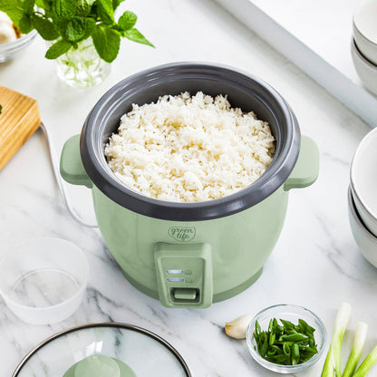 Sage green 6-cup rice cooker with ceramic nonstick pot, filled with rice on a kitchen counter near fresh herbs and utensils.