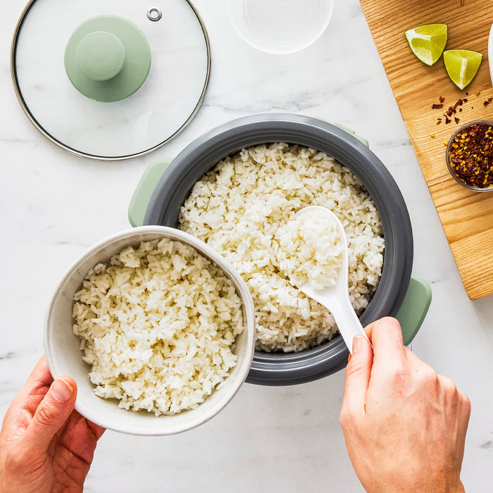 White rice being served from a sage green GreenLife 6-cup rice cooker into a bowl, with lime wedges and chili flakes nearby.