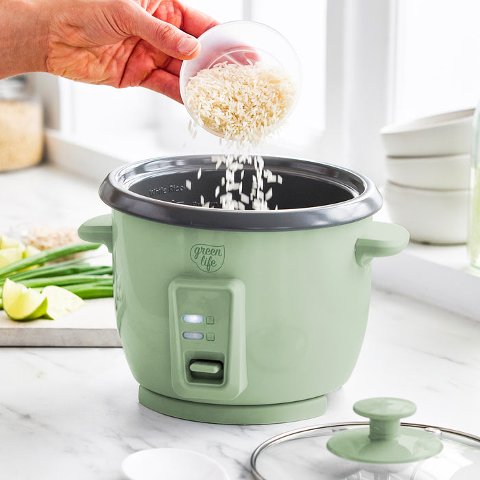 Hand pours rice into sage green GreenLife 6-cup rice cooker; fresh vegetables are on the kitchen counter nearby.