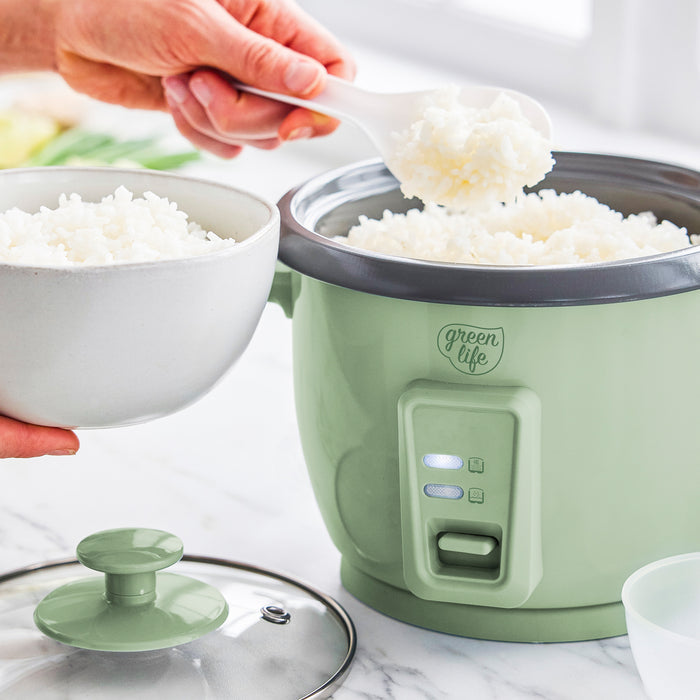 Hand scooping rice from sage green 6-cup GreenLife rice cooker into a white bowl on a kitchen counter.