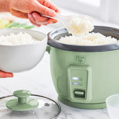 Hand scooping rice from sage green 6-cup GreenLife rice cooker into a white bowl on a kitchen counter.