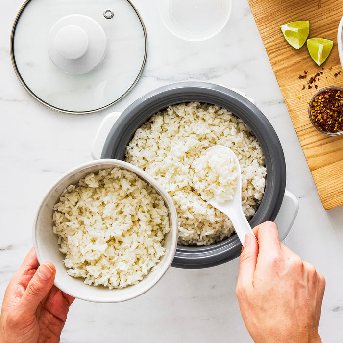 Rice being served from a white GreenLife 6-cup rice cooker into a bowl on a white countertop.