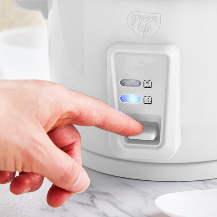 Hand pressing button on white GreenLife 6-cup rice cooker with two indicator lights on the front.