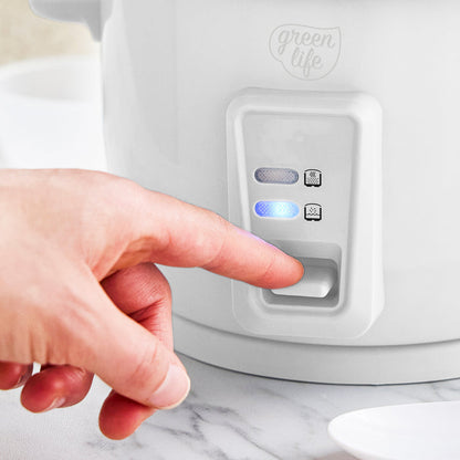 Hand pressing button on white GreenLife 6-cup rice cooker with two indicator lights on the front.