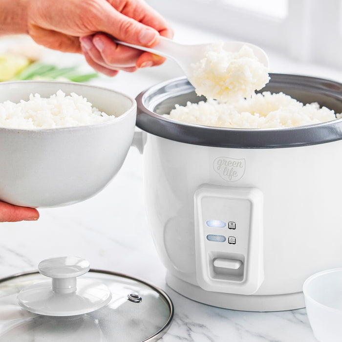 A hand scooping rice from a white GreenLife 6-cup rice cooker into a bowl on a white countertop.