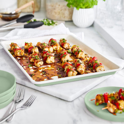 Chicken and vegetable skewers arranged on a sage green GreenLife ceramic nonstick baking sheet with herbs.