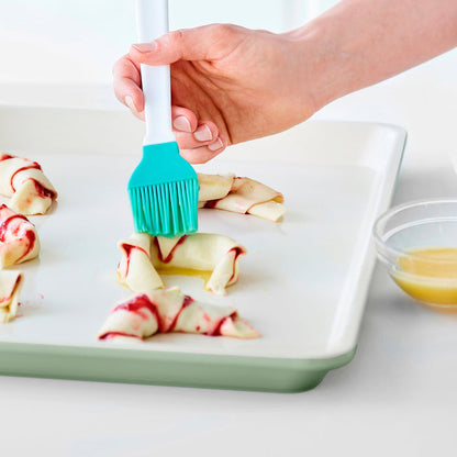 Hand brushing melted butter on pastry rolls atop a sage green ceramic nonstick cookie sheet.