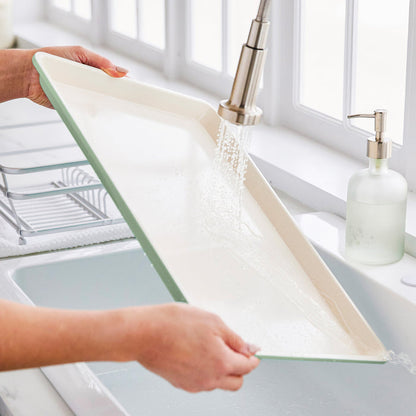Sage green ceramic nonstick cookie sheet being rinsed under running water in a kitchen sink.