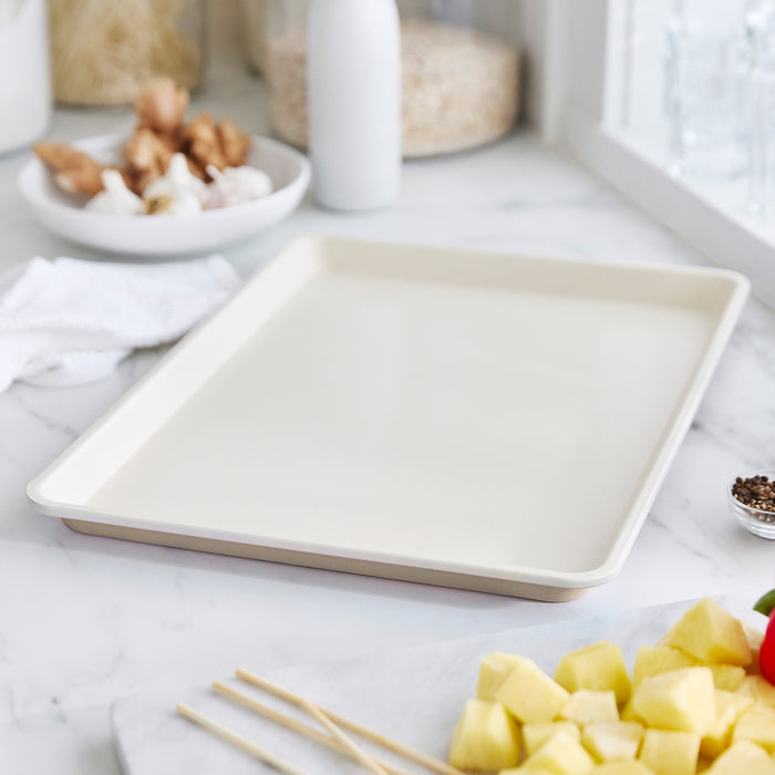 Taupe ceramic nonstick cookie sheet on a marble countertop with chopped vegetables and kitchen utensils nearby.