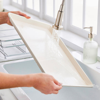 Person rinsing a GreenLife ceramic nonstick cookie sheet in a sunlit kitchen sink.