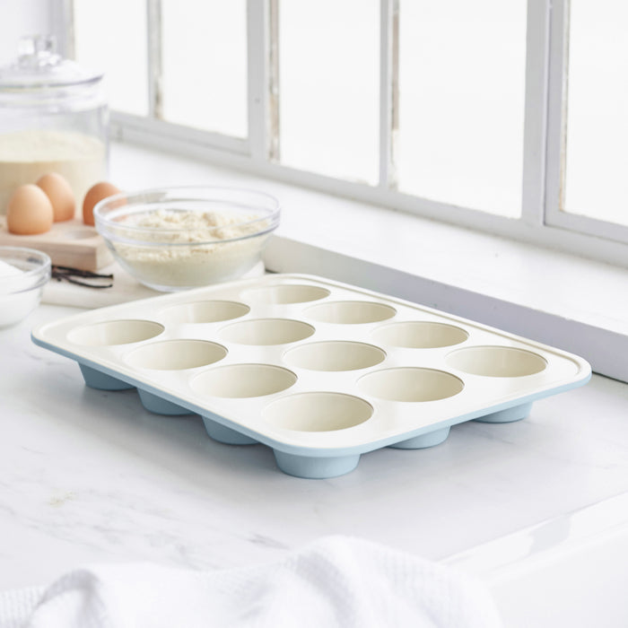 Blue ceramic nonstick muffin pan on a white countertop with baking items nearby in a bright kitchen.