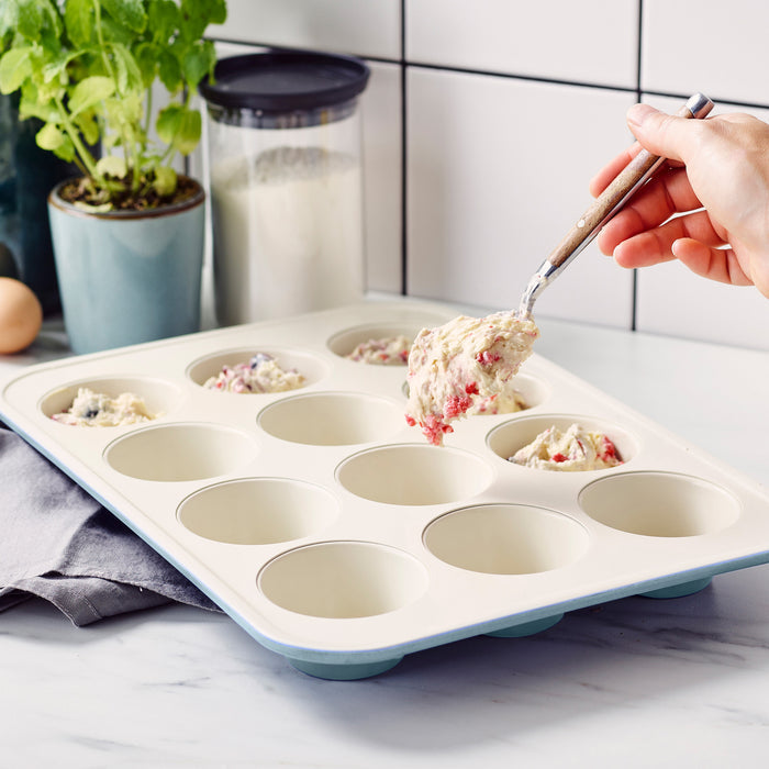 Hand spooning batter into a blue GreenLife ceramic nonstick muffin pan on a kitchen counter.