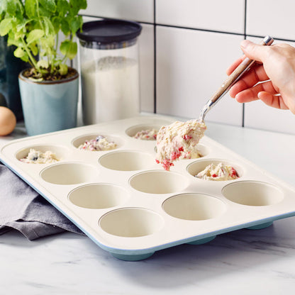 Hand spooning batter into a blue GreenLife ceramic nonstick muffin pan on a kitchen counter.