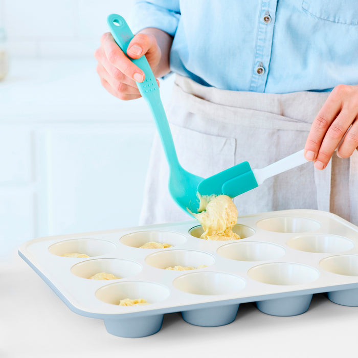 Batter being spooned into a blue GreenLife ceramic muffin pan with a spatula in a bright kitchen.