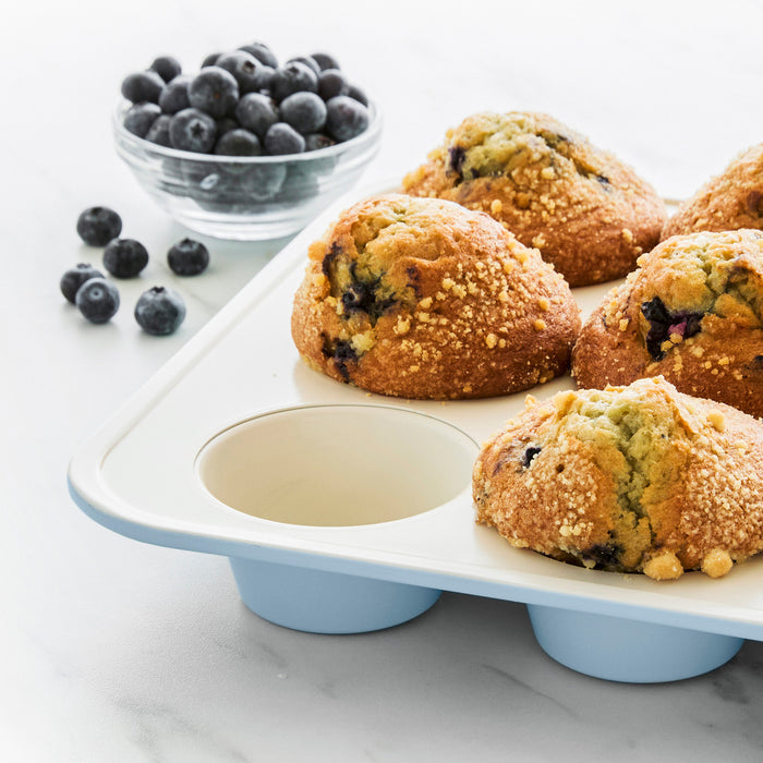 Blue ceramic muffin pan with five blueberry muffins, one slot empty, fresh blueberries in the background.