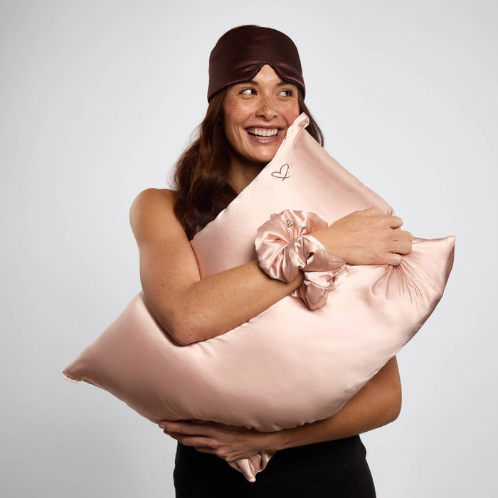 A Kitsch Nighttime Renewal 3pc Sleep Set with model with brown hair, holding in her arms a satin pillowcase, scrunchie on her wrist, and eye mask with heart details on her head.