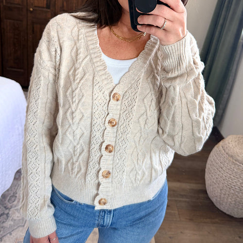 A woman wearing a cream cable-knit cardigan sweater with wooden buttons over a white top and blue jeans, taking a mirror selfie in a home setting.