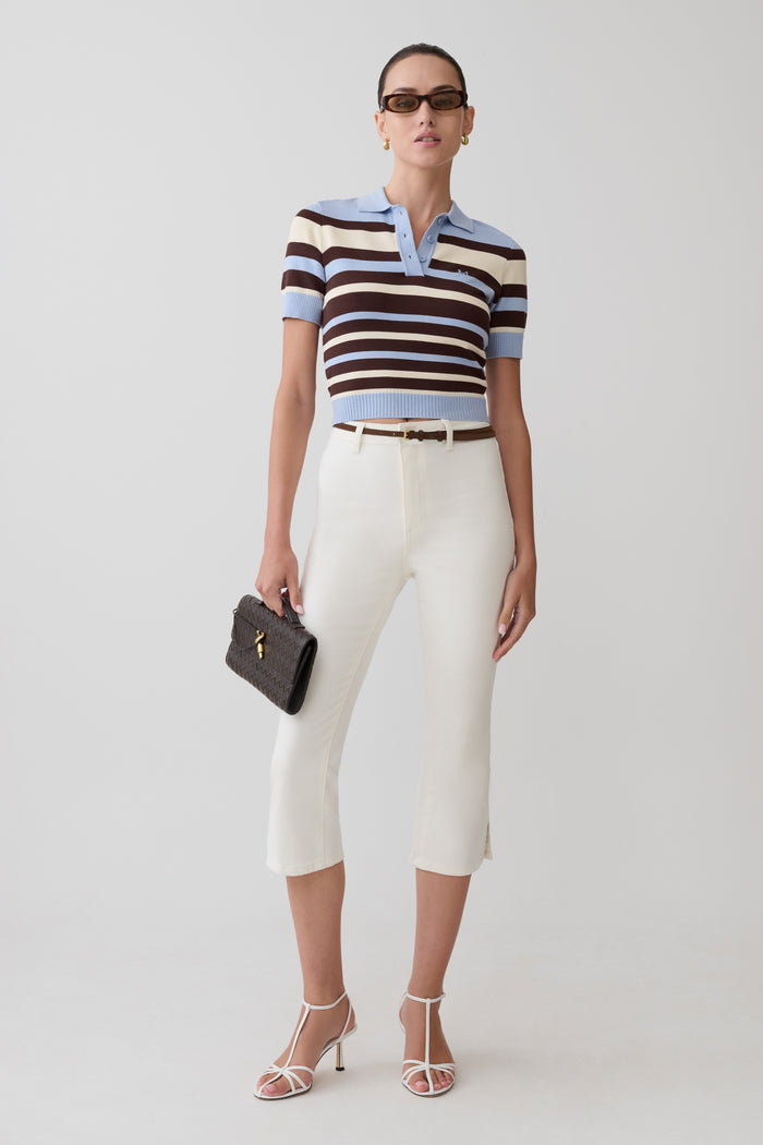 Woman wearing a striped polo shirt and white pants with a black clutch against a white background