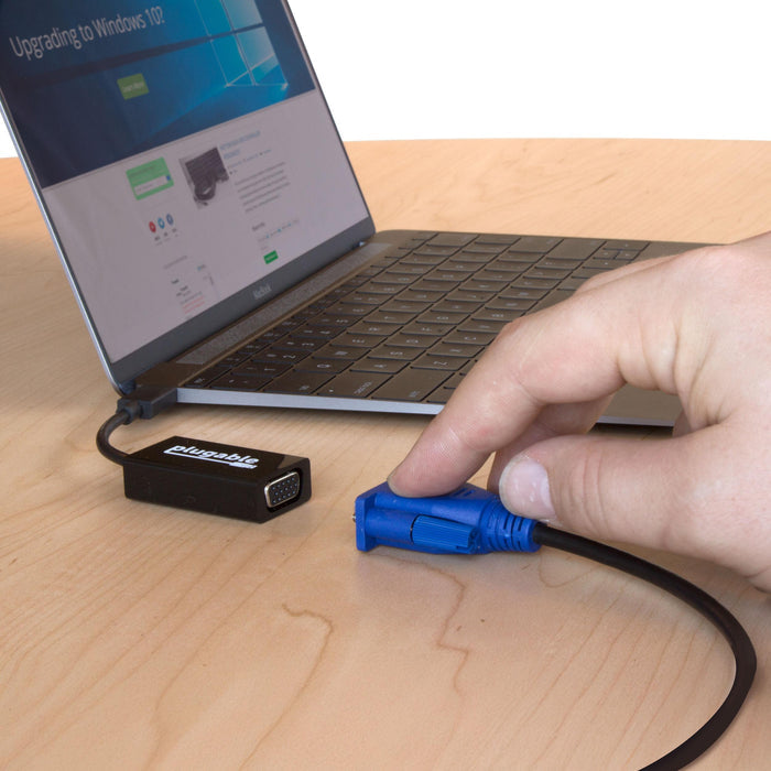 Plugable USBC-VGA adapter connected to a laptop, with a hand holding a VGA cable connector ready to plug in.