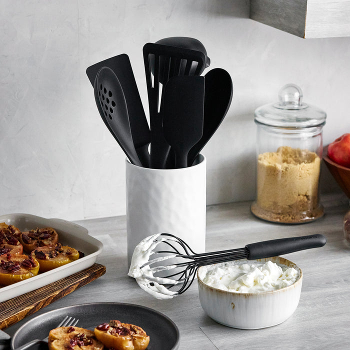 Black silicone utensil set with crock, eight pieces, on a kitchen counter beside a whisk and jar of brown sugar.