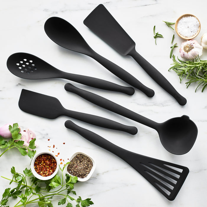 The Premiere Silicone Roasting Scoop in black, designed for durability, is displayed on a white marble surface with fresh herbs, garlic, and small bowls of salt, pepper, and red pepper flakes.
