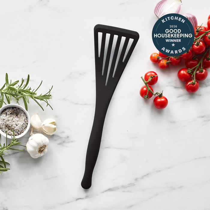 The Premiere Silicone Fish Turner | Black, a 2023 Good Housekeeping Kitchen Awards Winner, lies on marble among vine cherry tomatoes, rosemary, garlic, and seasonings—its durable and flexible design perfect for flipping fish.