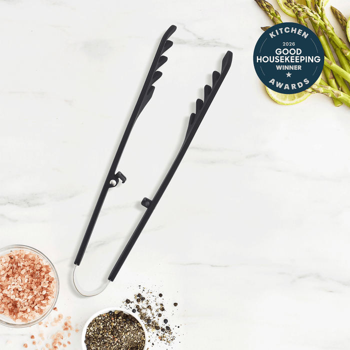 Premiere Silicone Locking Pasta Tongs | Black, featuring a steel core, rest on a white surface with pink salt, black pepper, asparagus spears, and a “Good Housekeeping 2023 Kitchen Awards Winner” badge in the corner.