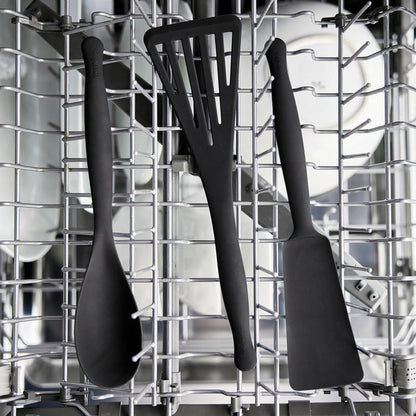 The Premiere Silicone Solid Spoon in black is shown upright in the dishwasher’s top rack with white dishes, demonstrating it is dishwasher safe.