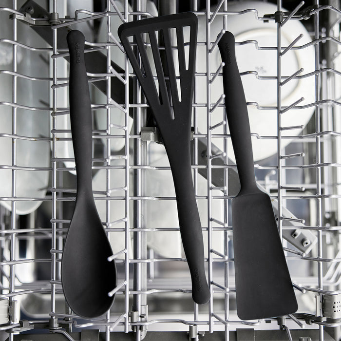 The Premiere Silicone Solid Spoon in black is shown upright in the dishwasher’s top rack with white dishes, demonstrating it is dishwasher safe.