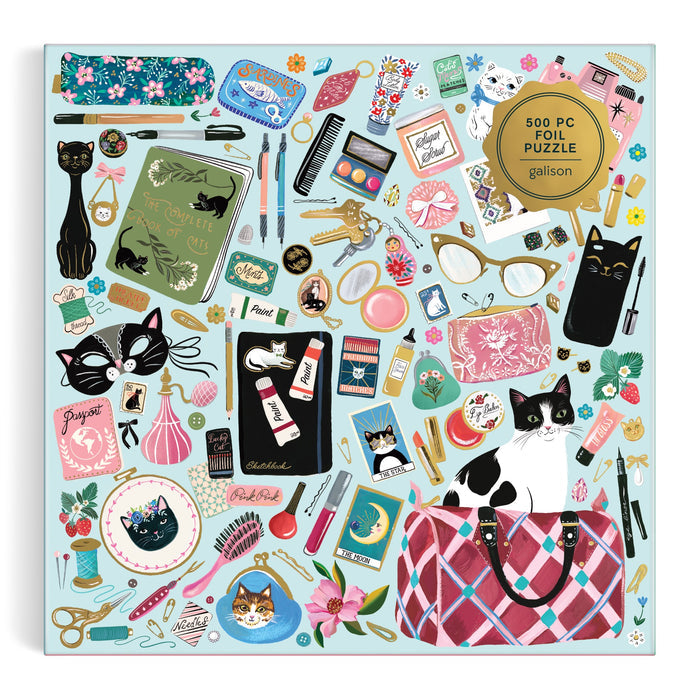 The Purrfect Accessories 500 Piece Foil Puzzle by Christine De Carvalho features illustrated cats, accessories, and stationery on a light blue background in a colorful, cat-themed display.