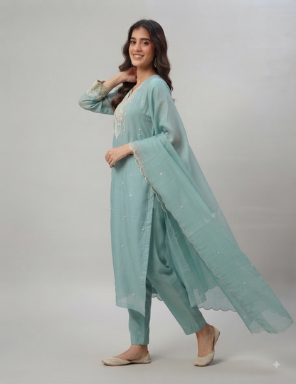 A woman modeling a light blue-green traditional outfit with matching dupatta, featuring embroidered details, against a plain light gray studio background.