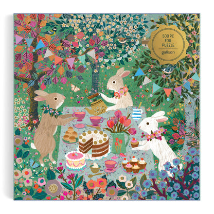 The Spring Tea Party 500 Piece Foil Puzzle by Victoria Ball features three rabbits at a whimsical outdoor tea party with cakes, teapots, flowers, and festive bunting amid lush greenery and trees.