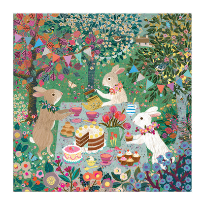 Three rabbits with flower garlands enjoy tea, cakes, and teacups at a festive table in a vibrant garden, surrounded by bunting and trees. Inspired by Victoria Ball art. Spring Tea Party 500 Piece Foil Puzzle by Victoria Ball.