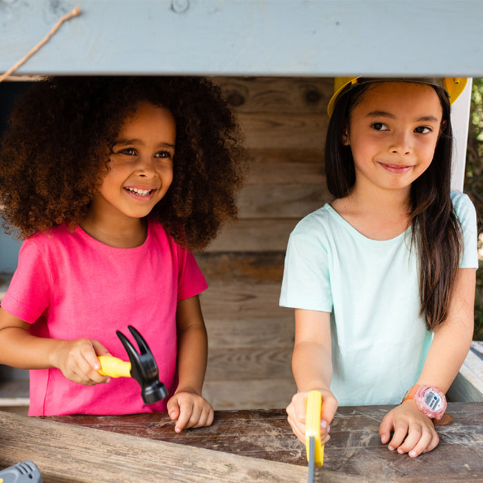 Two young girls stand at a workbench, smiling as one holds a hammer from the Red Toolbox Stanley Jr. 3 Piece Pretend Play Toolset and the other wears a yellow hard hat—both happily engaged in creative play.