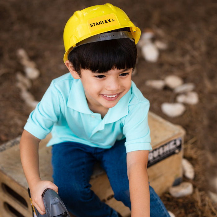 A young boy in a yellow construction helmet and light blue shirt sits on a wooden crate outdoors, smiling as he holds his Red Toolbox Stanley Jr. 3 Piece Pretend Play Toolset.