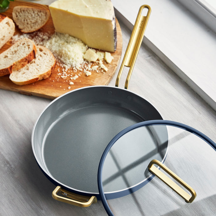 Blue ceramic nonstick frypan with lid next to a wooden cutting board holding bread and cheese.