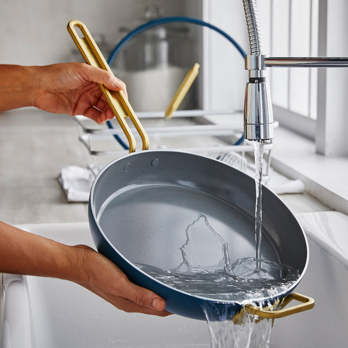 Washing a blue ceramic nonstick frying pan with lid under a kitchen faucet.
