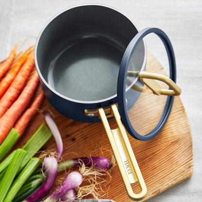 Blue ceramic nonstick saucepan with lid on a wooden board, next to carrots, celery, and onions.