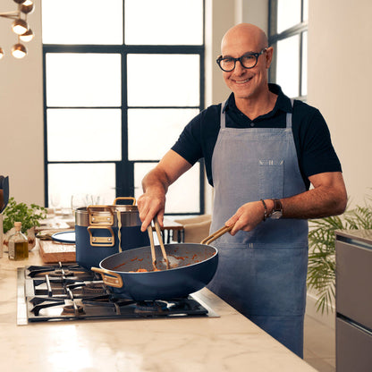 Man wearing glasses smiles while cooking with a blue Stanley Tucci 4.5-quart pan in a modern kitchen.