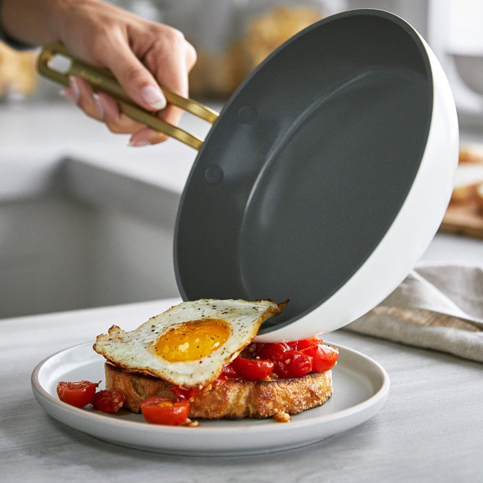 Hand serving a fried egg from an 8-inch white ceramic nonstick frypan onto toast with tomatoes.