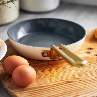 White ceramic nonstick 8-inch frypan with gold handle on a board next to two brown eggs.