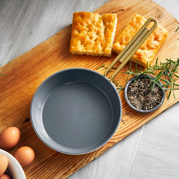 White ceramic frypan with bread, eggs, rosemary, and pepper beside it on a wooden surface.