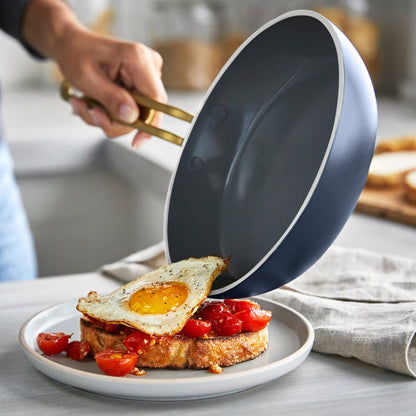 Hand sliding a fried egg from a blue ceramic nonstick 8-inch frypan onto toast and tomatoes.